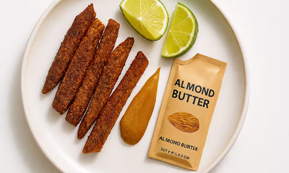 Close-up of a white plate holding six crispy, seasoned chili-lime seitan strips arranged in a row, a dollop of almond butter, a small beige packet labeled “Almond Butter” with an almond illustration, and two lime wedges. The setup is on a light background, shot in bright natural lighting.