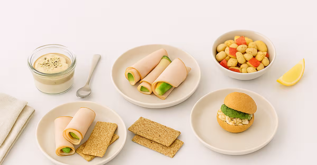 Flatlay of back-to-school high-protein breakfasts including a quark–tahini espresso date jar with hemp seeds, turkey–cheese avocado roll-ups with crackers, a lupini bean apple-cinnamon cup, and a canned-chicken pesto slider, arranged on neutral ceramic plates with a lemon wedge.