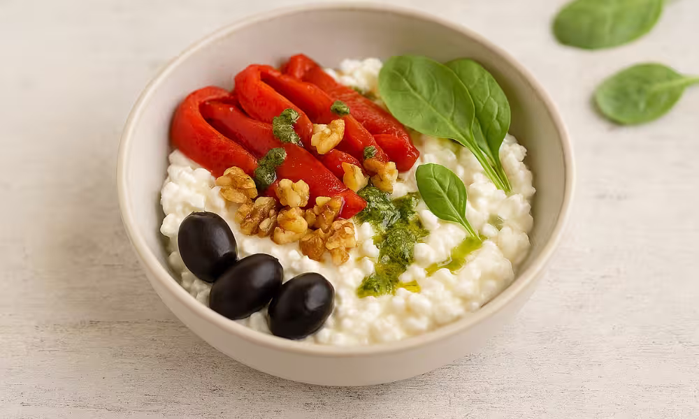 A rustic bowl of creamy cottage cheese topped with sliced roasted red peppers, crumbled walnuts, and a drizzle of fresh green basil pesto, served on a light wooden table with natural daylight.