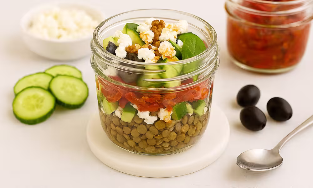 Cold lentil salad in a glass jar layered with lentils, goat cheese, cucumbers, olives, and sun-dried tomato oil.