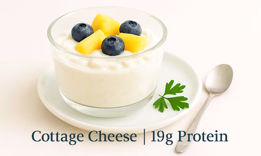 A glass bowl of creamy cottage cheese topped with fresh blueberries and pineapple chunks, placed on a white plate with a parsley garnish. A silver spoon rests beside the plate on a light surface. The image has bright, clean lighting with overlay text reading: “Cottage Cheese | 19g Protein.”