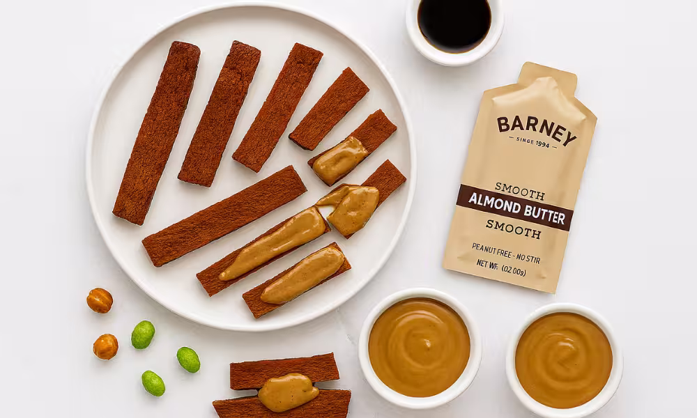 Overhead view of a white plate with brown seitan strips, some topped with smooth almond butter, alongside a beige almond butter pouch, two small bowls of almond butter, a small dish of soy sauce, and scattered roasted chickpeas and freeze-dried edamame on a white background.