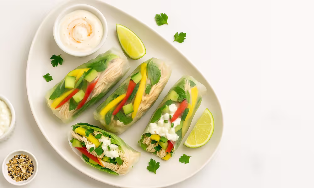 “No-cook mango-chili chicken rice-paper rolls with cucumber, bell pepper, mint, and a yogurt-chili dip on white marble.”