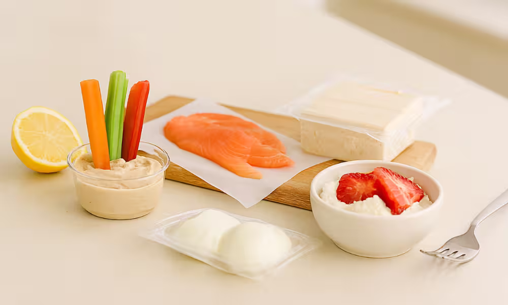 “Hummus cup with veggie sticks, smoked salmon slices, opened pack of tofu, and ricotta with strawberries on a bright kitchen counter.”