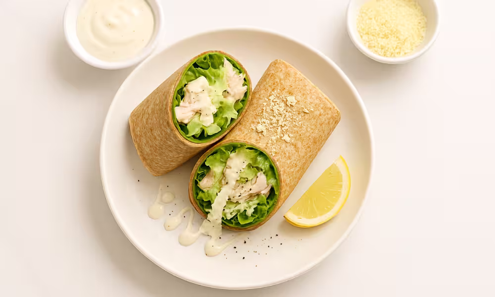 “High-protein chicken Caesar wrap sliced in half with romaine, yogurt Caesar dressing, parmesan and black pepper on white marble.”
