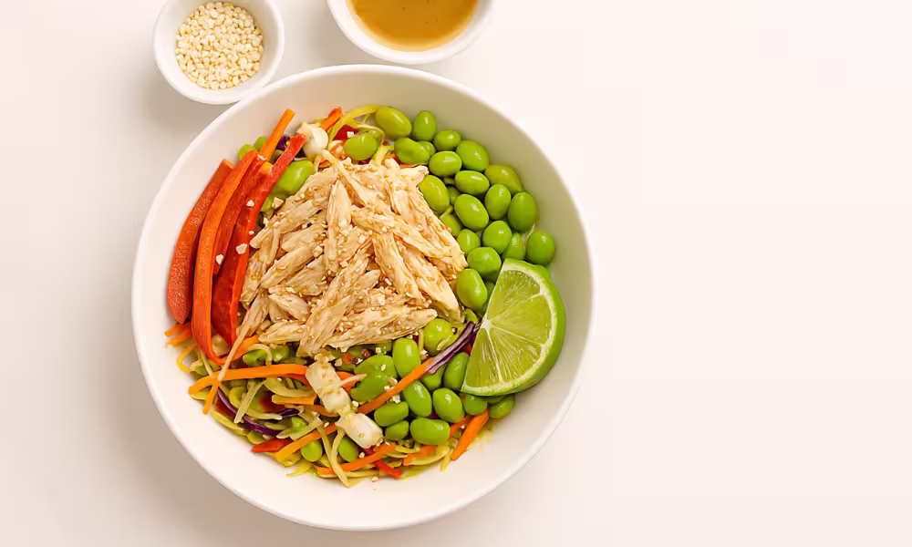 “Crunchy sesame chicken salad with coleslaw mix, edamame, red pepper, sesame seeds and lime on white marble.”