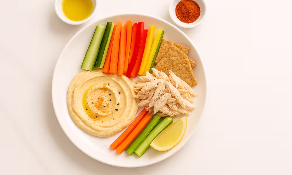 “Hummus & chicken rainbow plate with veg sticks, whole-grain crackers, olive-oil drizzle, paprika and lemon on white marble.”
