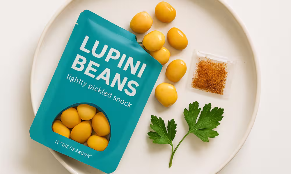 A turquoise pouch labeled “Lupini Beans – lightly pickled snack” sits on a white plate, surrounded by loose golden lupini beans, a small packet of reddish-brown seasoning, and two fresh parsley leaves.