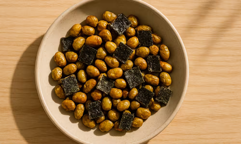 A bowl of roasted edamame beans mixed with small square pieces of dried seaweed, served in a beige ceramic dish on a light wooden surface.